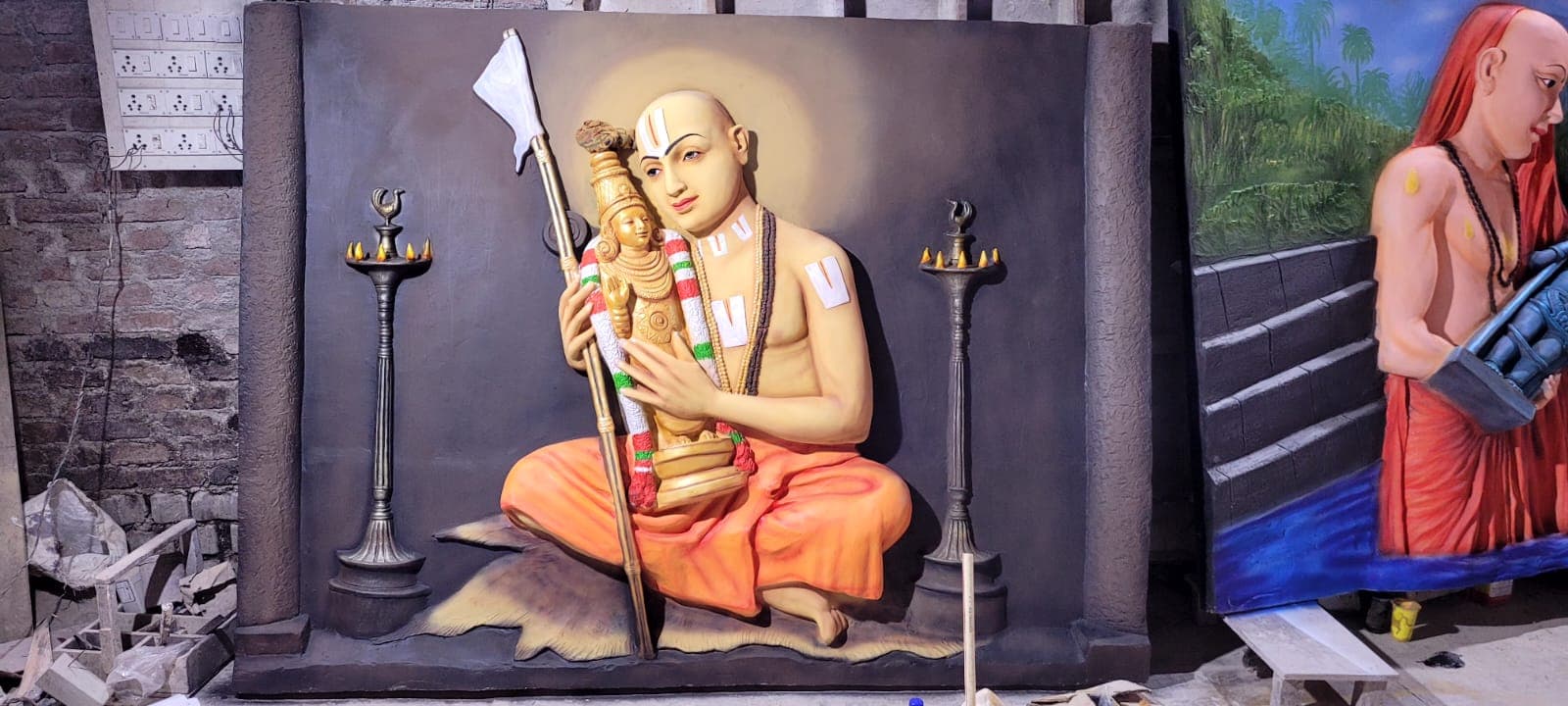 FRP relief panel of a seated acharya holding a deity beside temple oil lamps