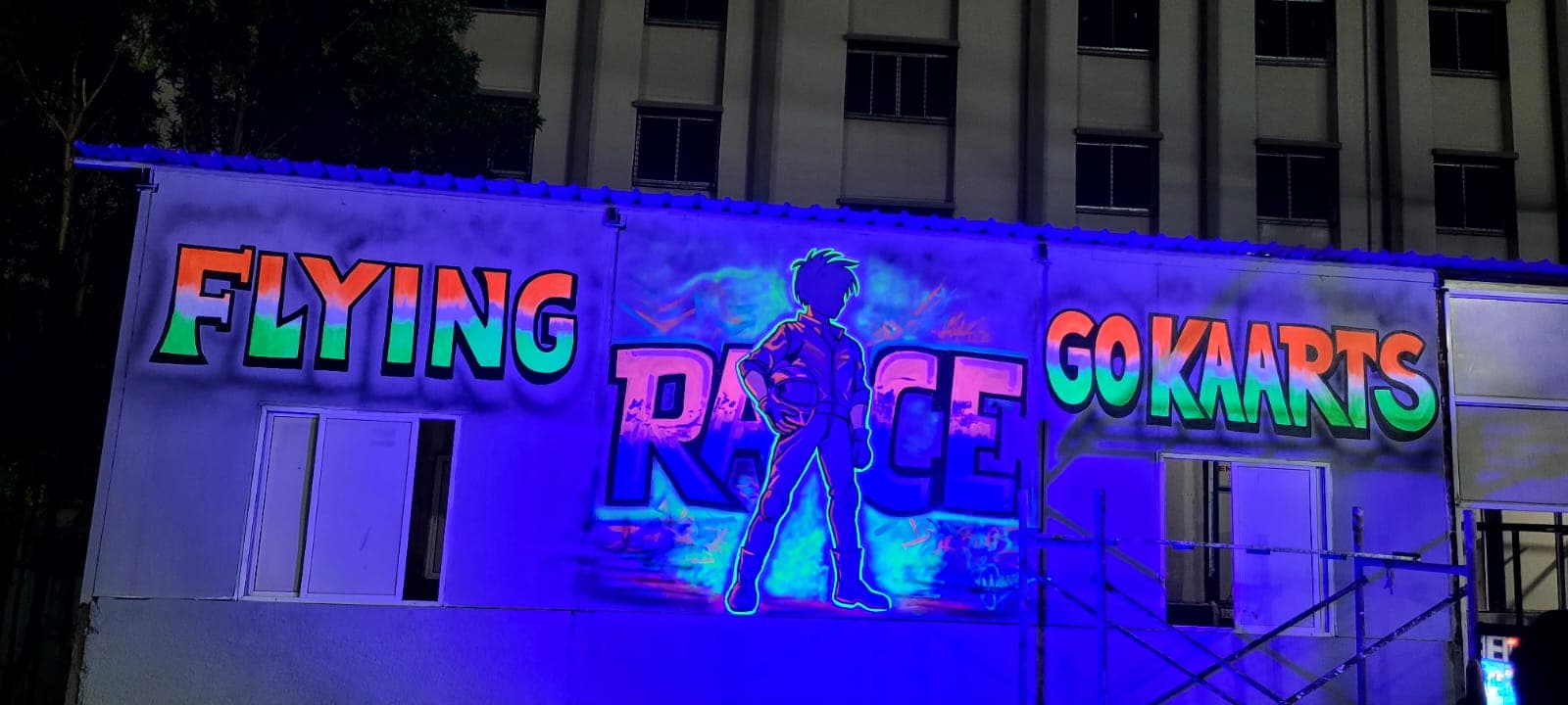 UV-reactive neon mural across the Flying Race Go Kaarts facade, photographed at night under blacklight — glowing lettering and a racing driver figure